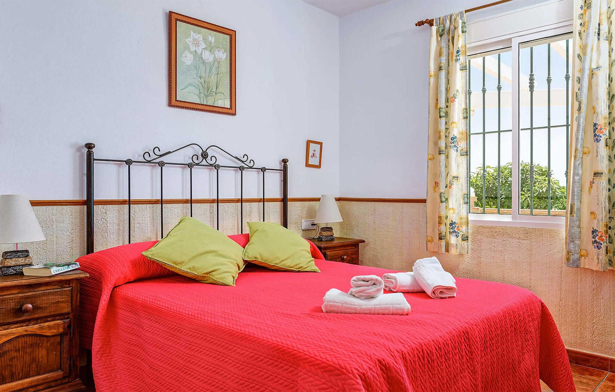 A bedroom with a red bedspread, green pillows, a framed picture on the wall, and a window with floral curtains.