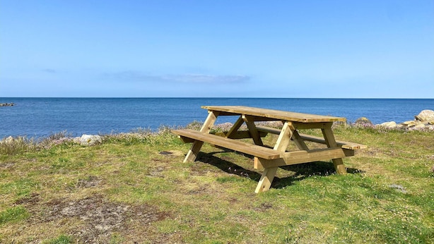 Una mesa de picnic de madera con bancos, ubicada en un área verde con vista al mar.