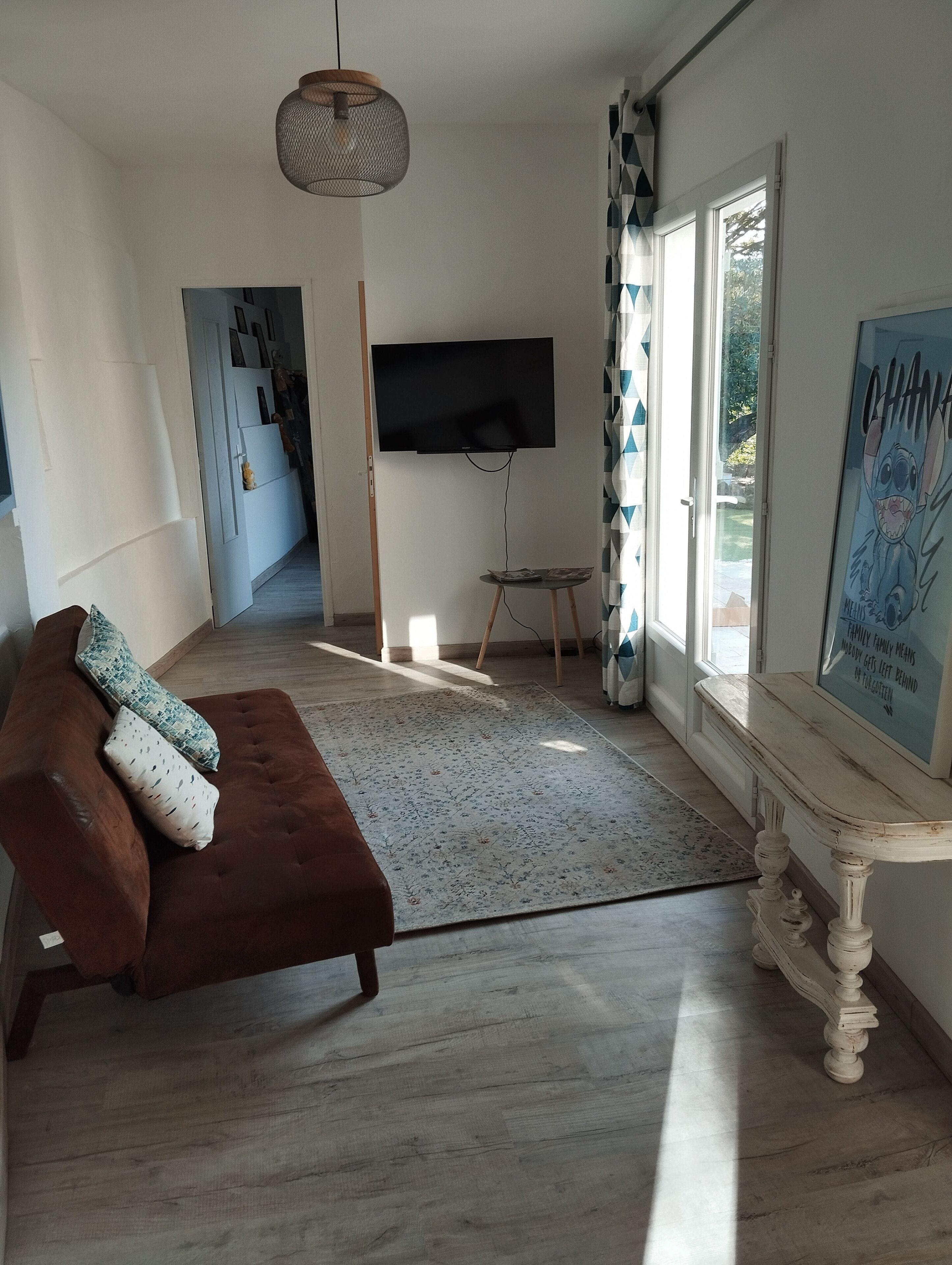 Een moderne woonkamer met een bruine bank, een flatscreen televisie aan de muur, een houten salontafel en een groot raam met uitzicht op de omgeving.