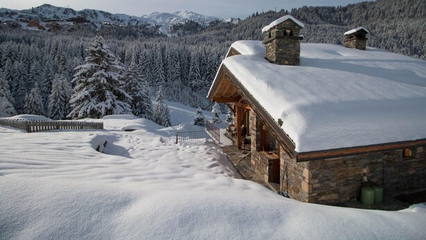 Eine steinerne Hütte mit schneebedecktem Dach, umgeben von verschneiter Landschaft und dichtem Wald.