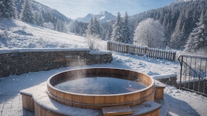 Ein hölzernes Whirlpoolbad, umgeben von einer Steinsäule, in einer verschneiten Winterlandschaft mit Bergen im Hintergrund.