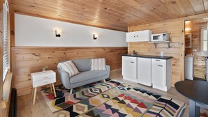 Ein kompakter, moderner Raum mit Holzwänden und -decke, einem grauen Sofa mit Kissen, einem kleinen weißen Nachttisch, einem weißen Schrank, einer Mikrowelle und einem gemusterten Teppich.