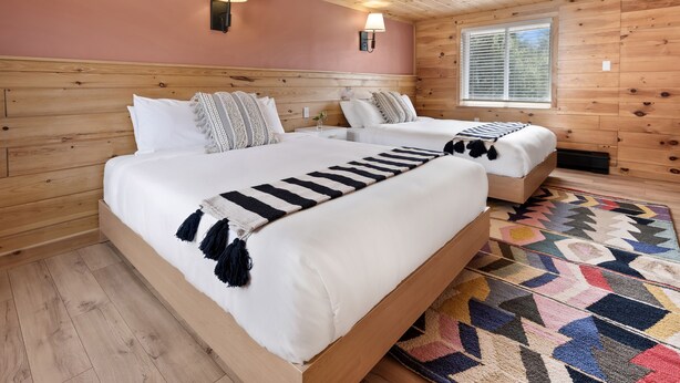 Ein Schlafzimmer einer Holzhütte mit zwei Betten, einem bunten Teppich und einem Fenster mit Jalousien.