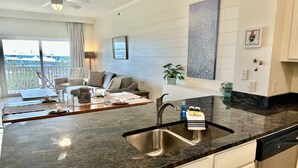 Eine moderne Küche mit Granitabdeckung, Edelstahlspüle und weißen Schränken. Es gibt einen Essbereich mit einer Couch und Blick ins Freie.