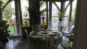 A covered patio with a round table set for dining, surrounded by white chairs, and a large potted plant in the background. - Johnstone Mansion (Keokuk)