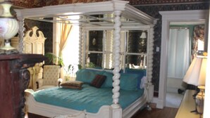 A four-poster canopy bed with a blue bedspread and pillows, a wooden chair, and a lamp on a table. - Johnstone Mansion (Keokuk)