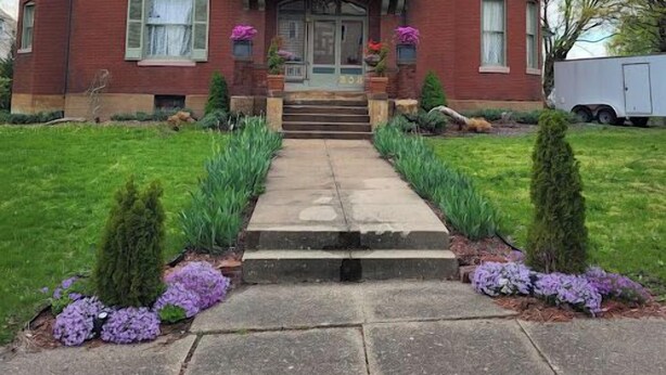 A large, two-story brick house with a prominent dome on the right side, a front porch with steps, and a well-maintained front yard with purple flowers and green shrubs. - Johnstone Mansion (Keokuk)