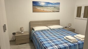 Een slaapkamer met een bed, twee nachtkastjes, een lamp, een schilderij van een strand en een raam met gordijnen.