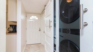 Un pasillo estrecho con una puerta blanca al final, una cafetera sobre una encimera y dos electrodomésticos negros, posiblemente una lavadora y secadora, a la derecha.