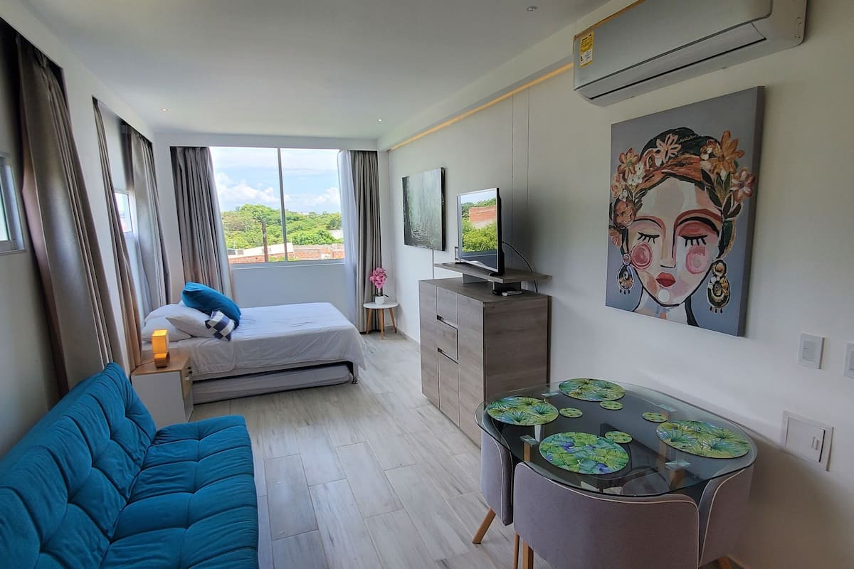 A modern hotel room with a bed, a blue sofa, a small table with decorative plates, a TV, and a large painting on the wall.