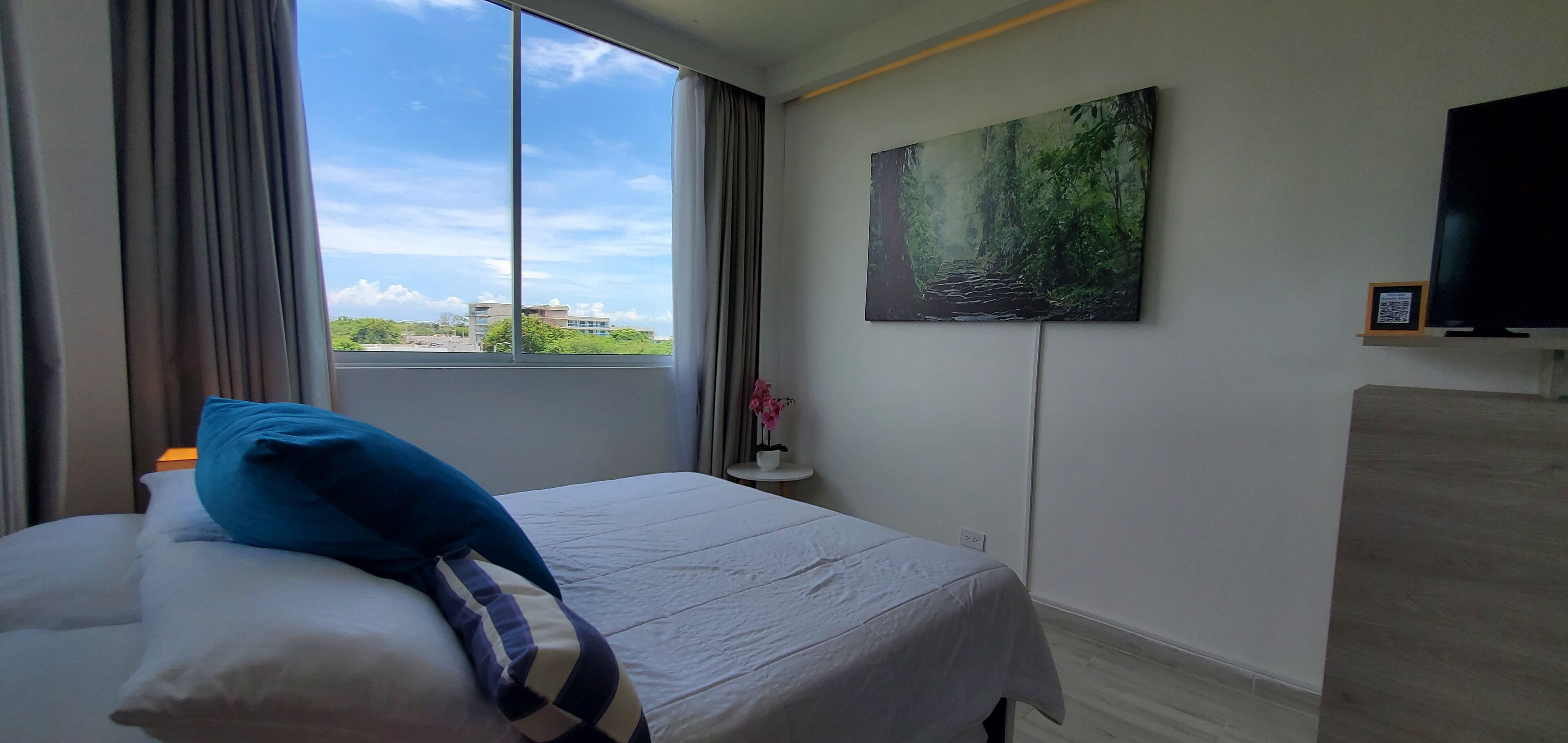 A hotel room with a large window, a bed with a blue pillow, a painting of a forest, and a TV on the wall.