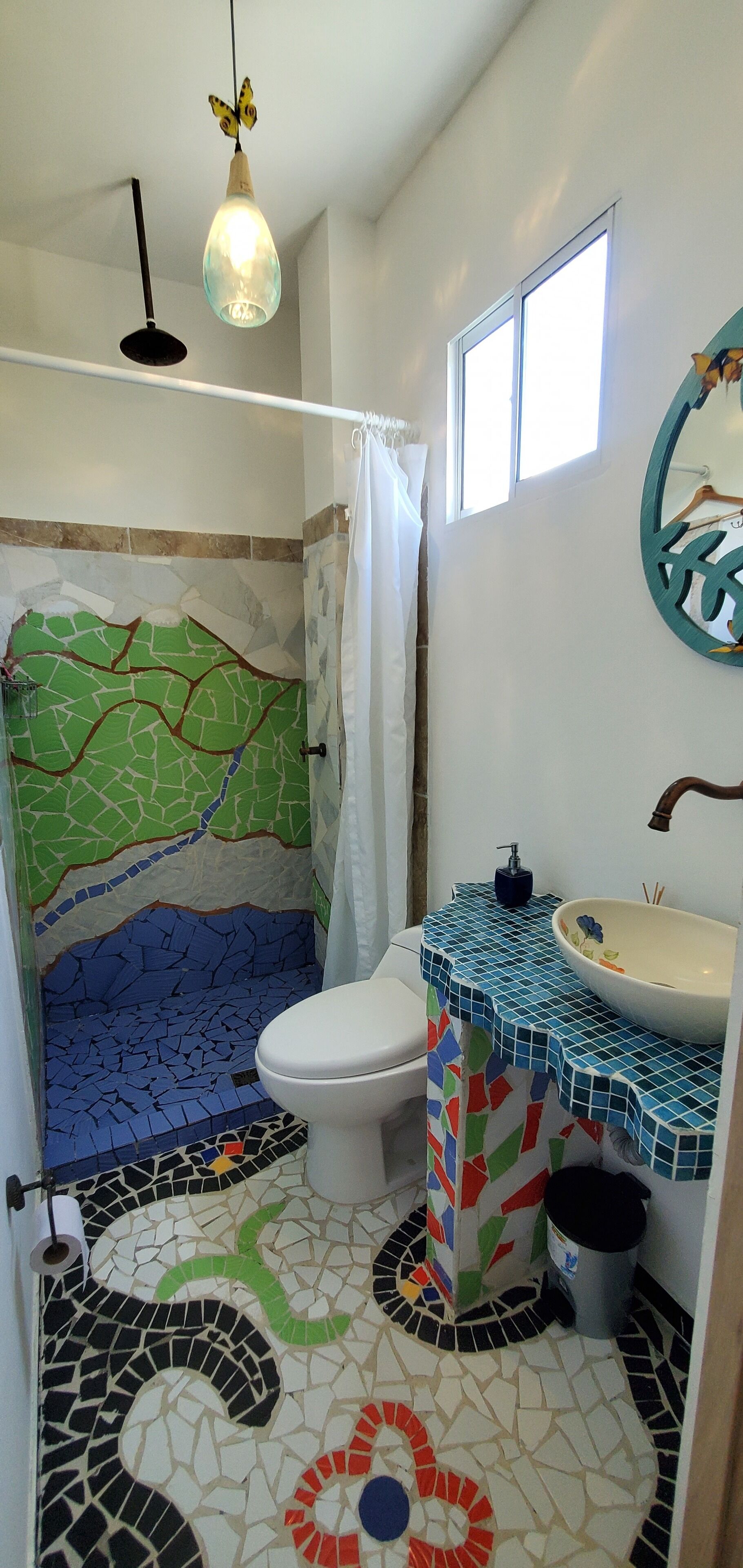 A bathroom with a mosaic floor, a toilet, a sink with a decorative basin, a hanging light fixture, and a window.