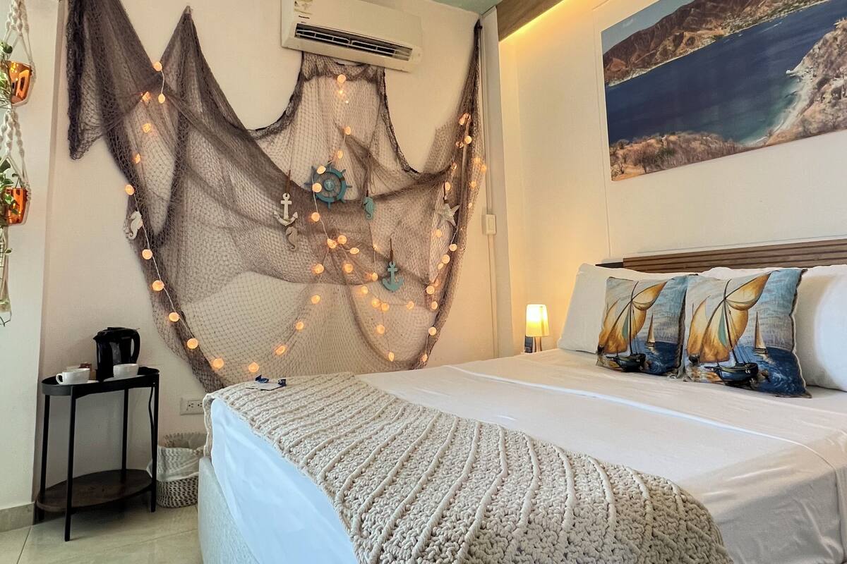 A bedroom with a bed, a wall-mounted air conditioner, a framed picture, a bedside table, and a lamp.