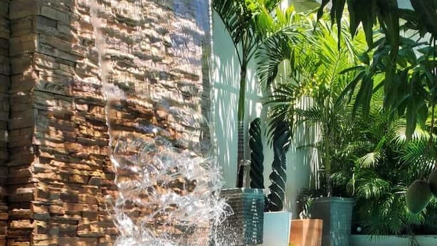 Une piscine agrémentée d’une cascade, entourée d’une végétation luxuriante et d’un mur de pierre.