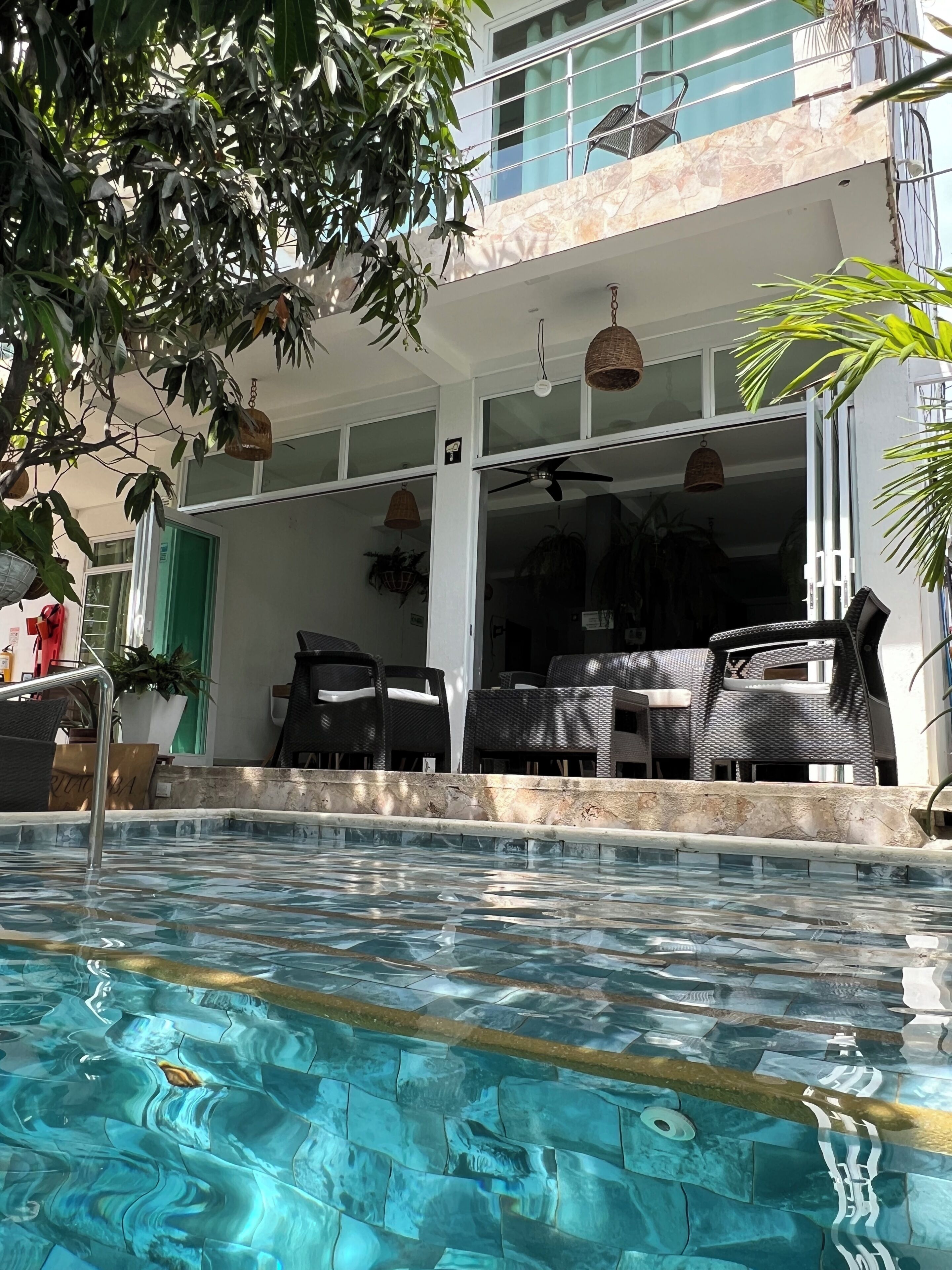 Une piscine avec un bord carrelé, un espace extérieur aménagé avec des meubles en osier, et un bâtiment doté d’un balcon.