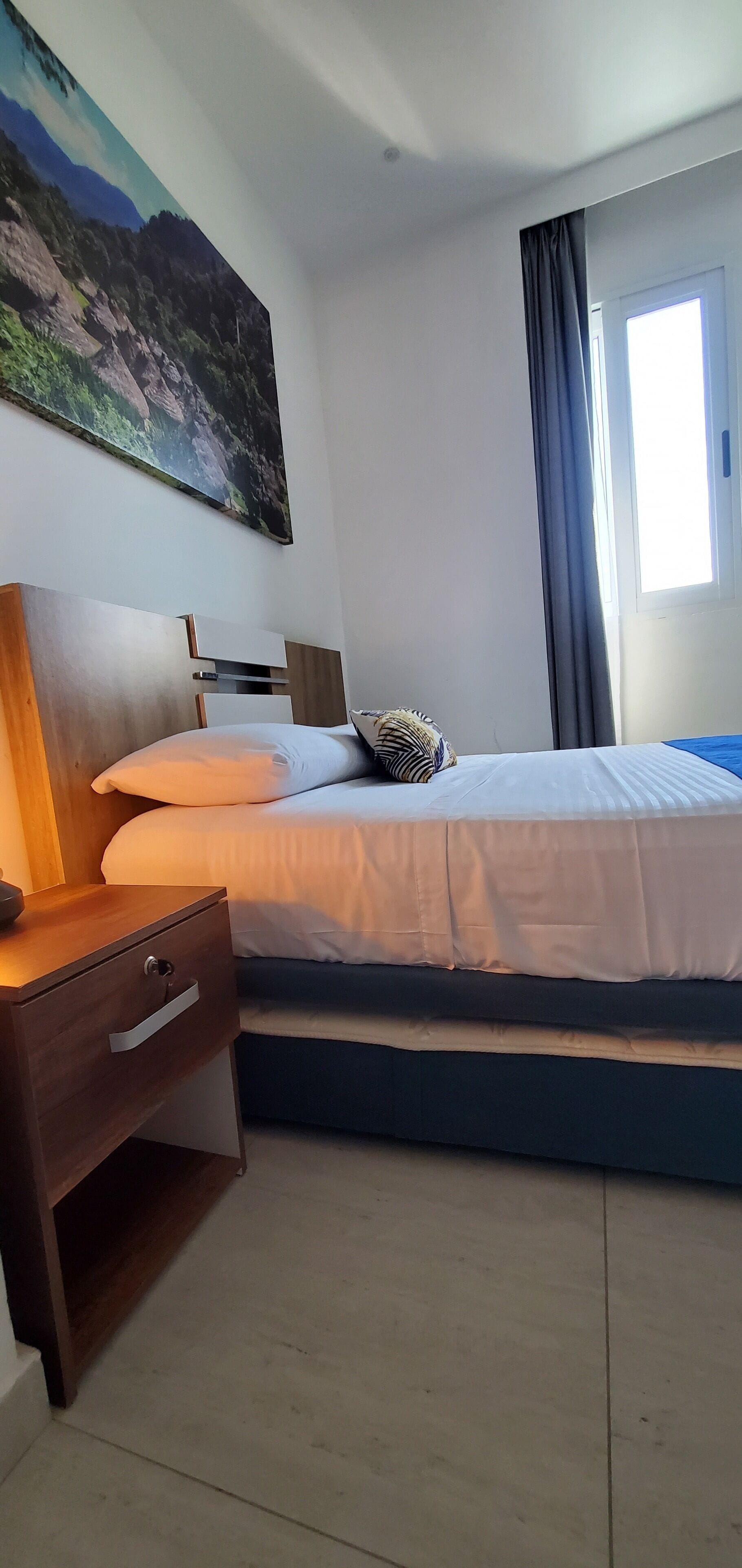 Ein Schlafzimmer mit Bett, Nachttisch, Lampe, einem Fenster mit Vorhängen und einem Bild an der Wand.