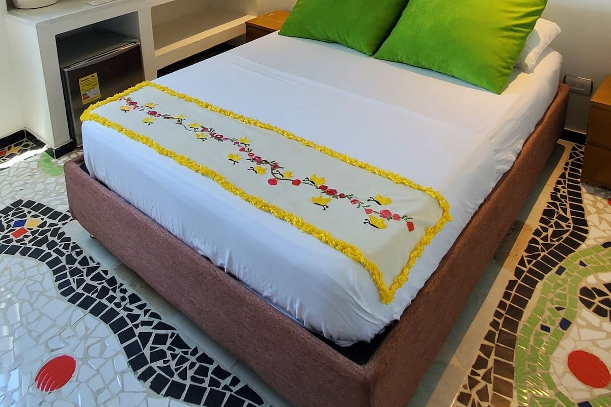 A neatly made bed with a white and yellow floral bedspread, green pillows, and a brown headboard. A large window with a view of a tree and landscape is visible.