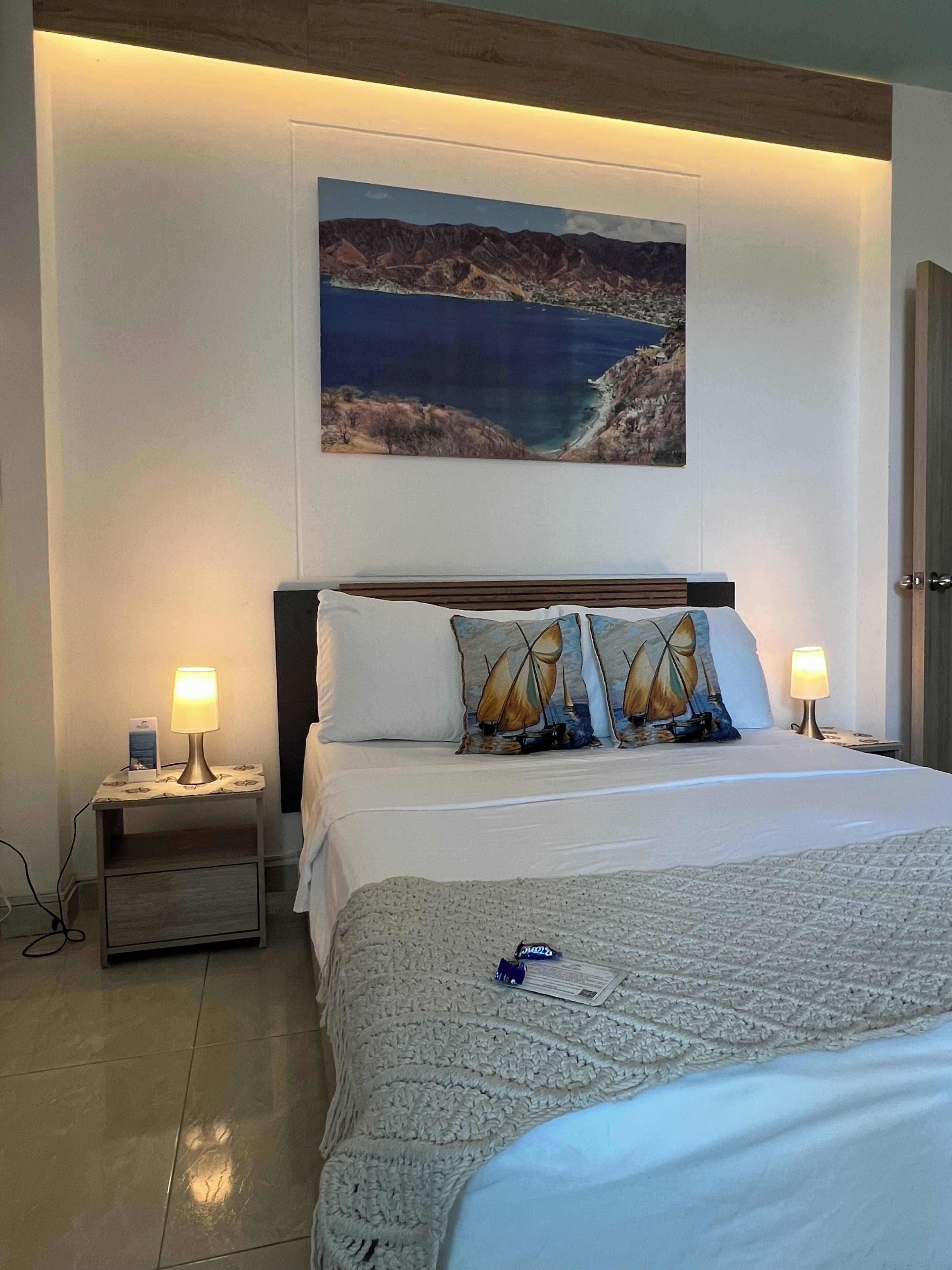 A bedroom with a bed, two bedside tables, a nightstand, a wall-mounted painting, and a door.
