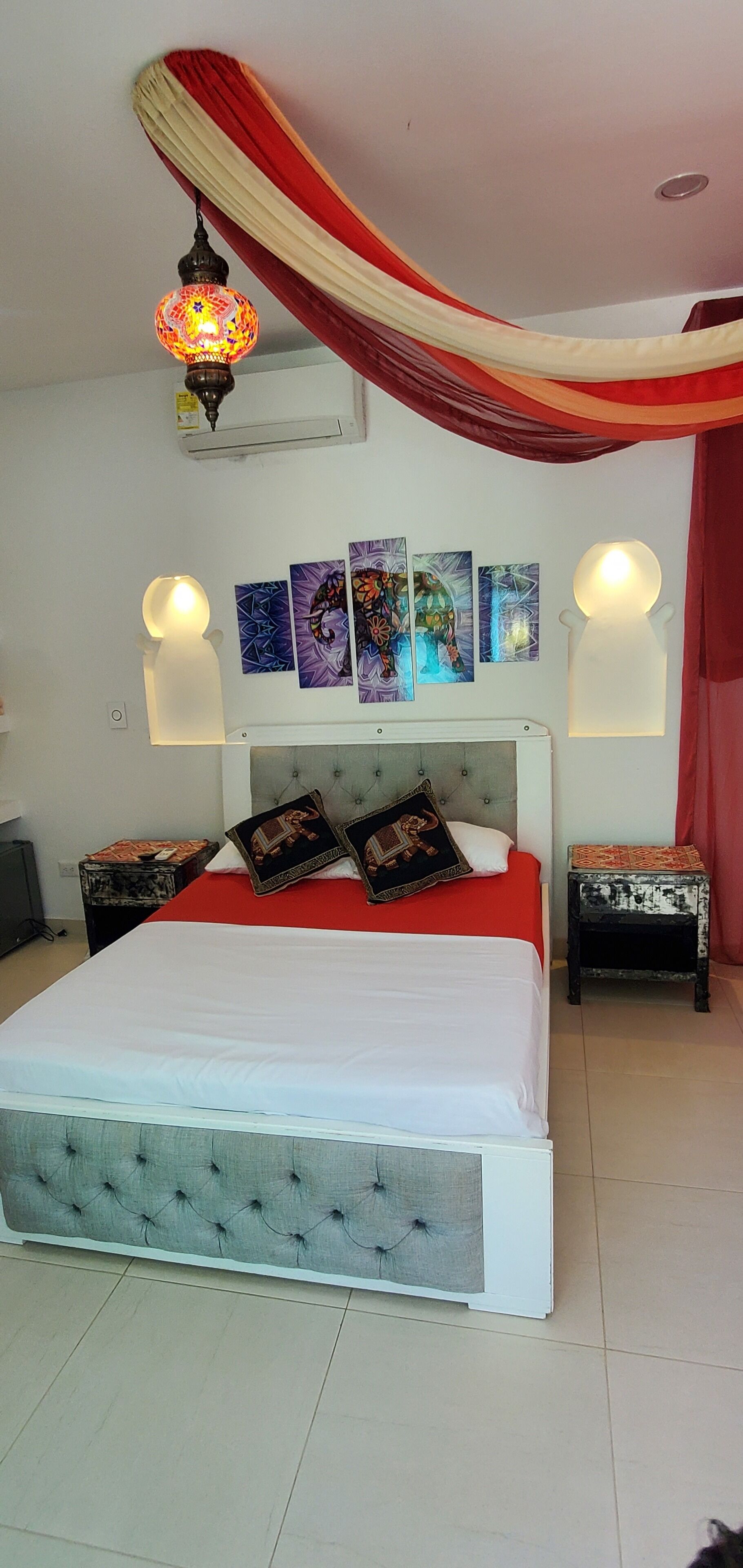 Ein Schlafzimmer mit einem großen Bett, einer gepolsterten Kopfteil, einer rot-weißen, drapierten Decke und Wandleuchten.