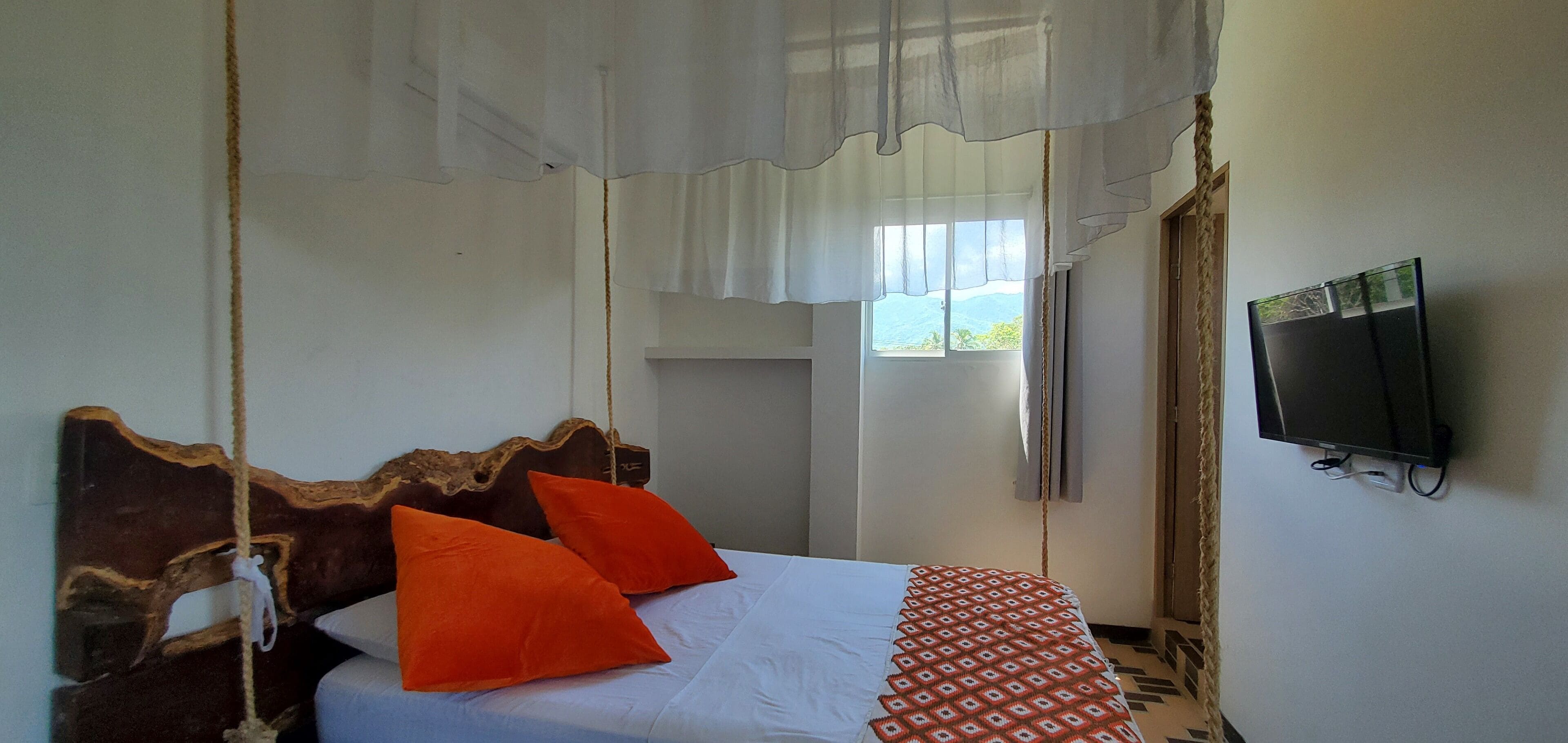 Quarto com cama, duas almofadas laranja, colcha branca, televisão na parede e janela com vista.