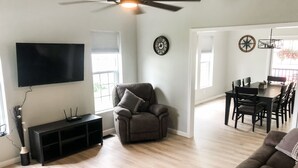 A living room with a flat-screen TV, a ceiling fan, a sofa, a dining table with chairs, and a small potted plant.