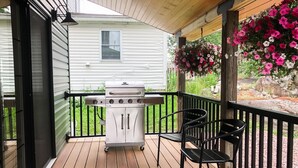 A covered outdoor deck with a grill, chairs, and hanging flowers.