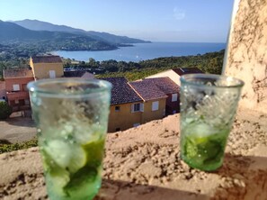 Two glasses of a green beverage on a balcony overlooking a coastal landscape with buildings and mountains in the distance.