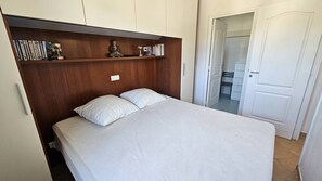 Un dormitorio con una cama, cabecera de madera, estante con libros y una estatua, y una puerta que da al baño.