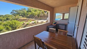 Un balcón con mesa y sillas de madera, una ventana con vistas a zonas verdes y un vehículo, y una puerta corrediza.