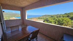 A balcony with a wooden table and chairs, offering a view of a coastal landscape with hills and a body of water. - Appartement SARAH Vue mer- Clim- Wi-fi (FAVONE CONCA)