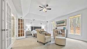 A spacious room with a high ceiling, a ceiling fan, a large window with a view of trees, and a door leading to another room.