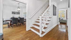 A staircase with wooden steps and white railings, a dining area with a table and chairs, and a view of the outdoors through a window.