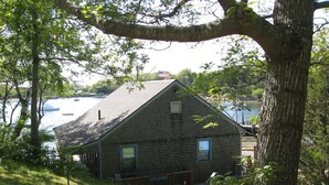 A rustic cabin with a sloped roof, situated on a grassy hill overlooking a body of water with boats. - Waterfront Boathouse Cottage (Falmouth)
