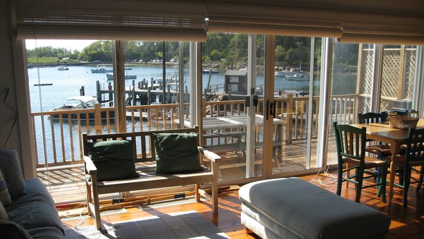 A room with a large window overlooking a dock with boats, a wooden deck, and a view of the water. There is a sofa and a chair with cushions. - Waterfront Boathouse Cottage (Falmouth)