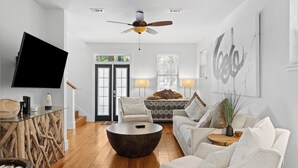Un salon spacieux au design moderne, comprenant une grande table basse ronde, un canapé, un téléviseur posé sur une table en verre et un ventilateur de plafond.
