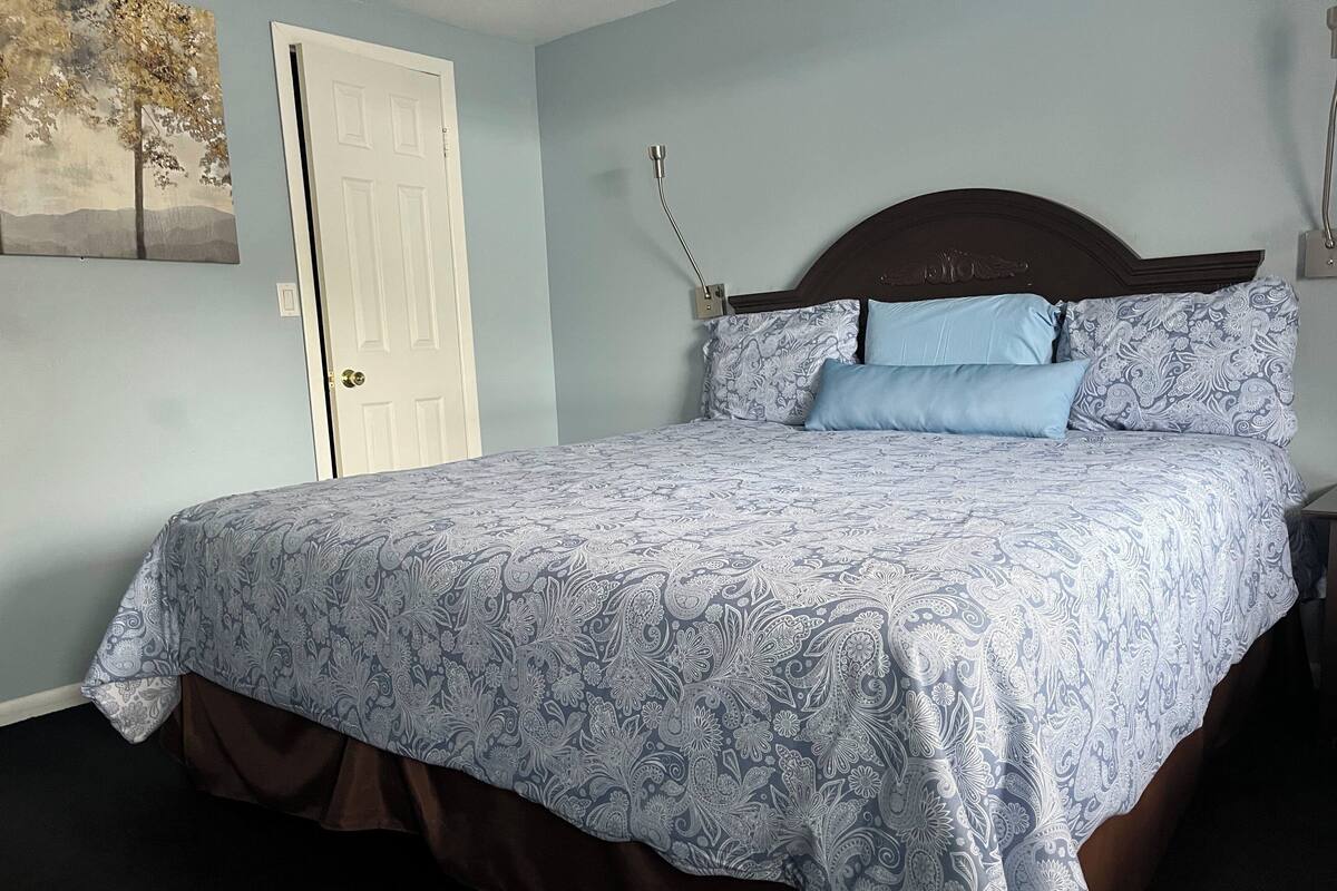 Quarto com cama, mesa de cabeceira, luminária na parede e porta.