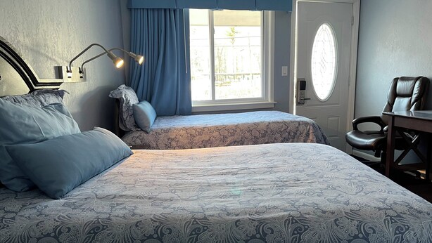 A bedroom with two beds, a desk, a chair, a window with curtains, and a door with a glass panel. - Hitching Post Country Motel (Cornwall Bridge)