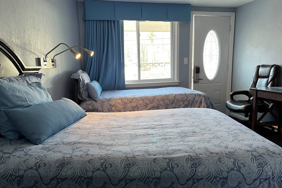 Quarto com duas camas, uma escrivaninha, uma cadeira, uma janela com cortinas e uma porta com painel de vidro.