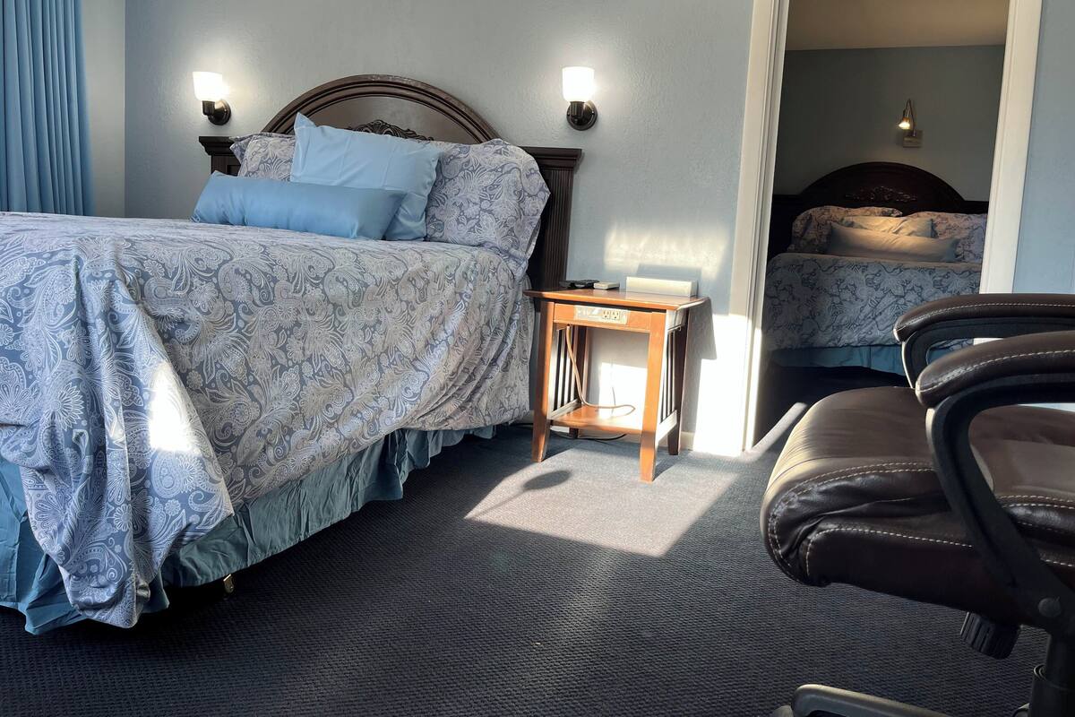 Quarto de hotel com uma cama grande, uma escrivaninha, uma cadeira e outra cama ao fundo.