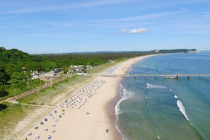 Ein Strand mit Reihen von Strandsonnenschirmen und Liegen, eine Mole, die ins Wasser führt, und ein Gebäudekomplex mit einem Garten.