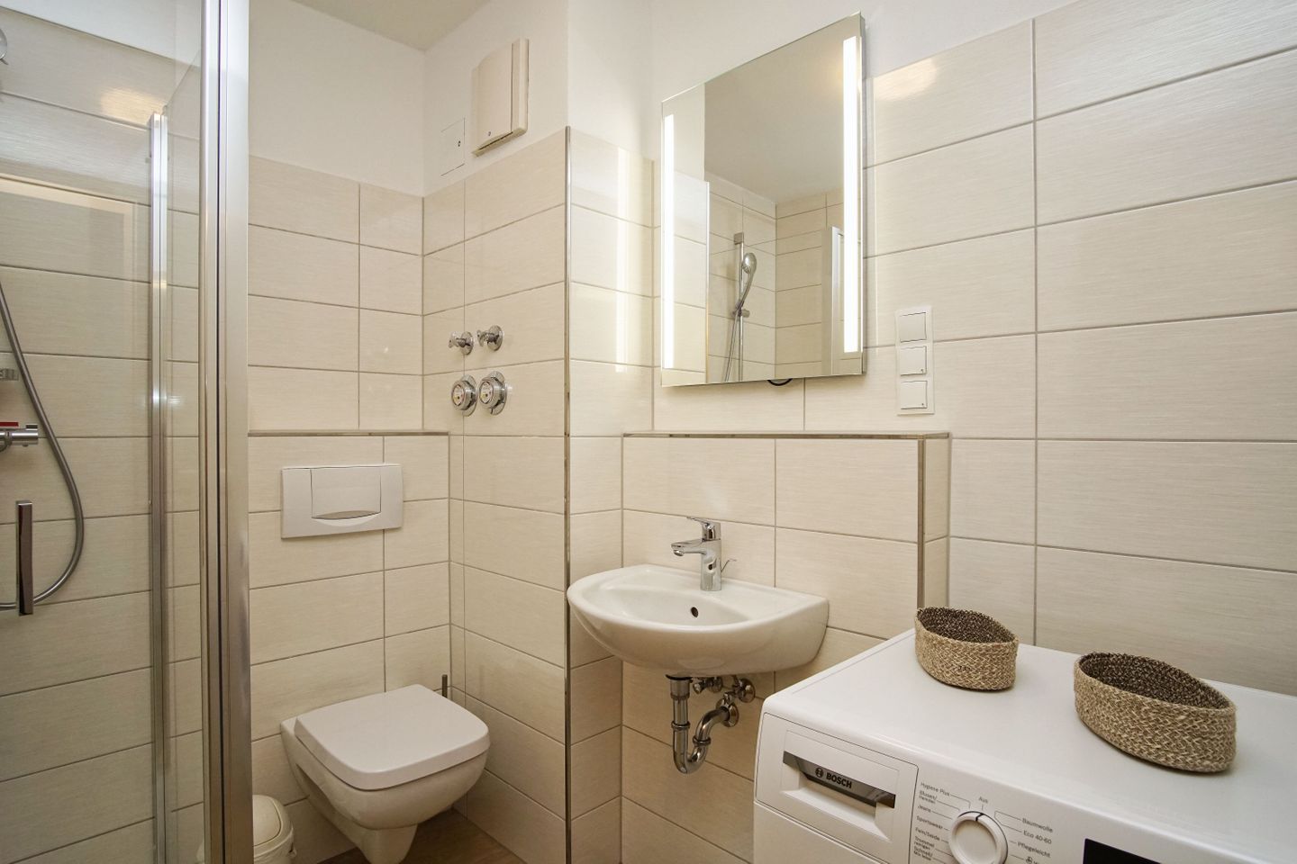 A bathroom with a glass shower enclosure, a toilet, a sink with a Bosch washing machine, and a mirror above the sink.