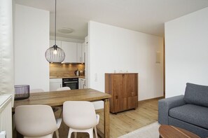 A modern kitchen with white cabinets, a wooden countertop, and a hanging light fixture. There is a dining table with two chairs and a grey sofa in the living area.