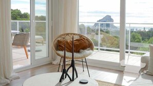 Habitació amb una finestra gran, una cadira de vimetera amb coixí, una taula rodona petita i vistes a una muntanya i un paisatge urbà.