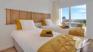 Habitació d'hotel amb dos llits, un capçal de llit de bambú i una finestra gran que ofereix vistes al mar i a la ciutat.