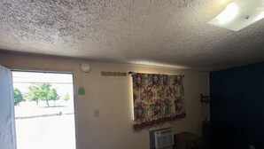 A room with a door leading to a parking lot, a wall-mounted air conditioner, a small trash bin, and a patterned curtain.