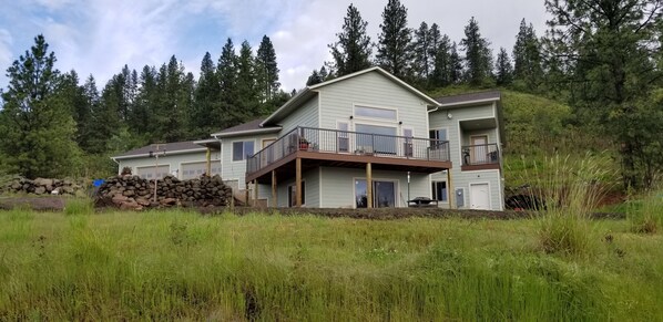 A two-story house with a balcony, surrounded by tall trees and a grassy field.