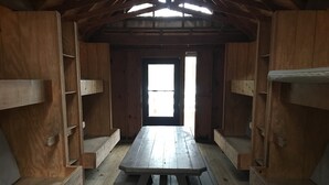 Une cabane en bois avec une table et des bancs au centre, une grande fenêtre et une porte.