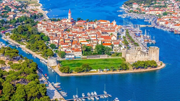 Vue aérienne d’une ville côtière avec un château historique, une marina où sont amarrés de nombreux bateaux et un ensemble dense de bâtiments aux toits rouges.
