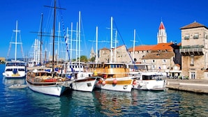Un port de plaisance où sont amarrés plusieurs bateaux, un bâtiment historique surmonté d’un toit de tuiles rouges et un ciel bleu azur.