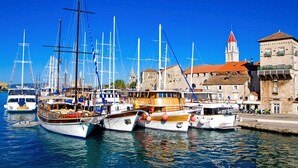 Un port de plaisance où sont amarrés plusieurs bateaux, un bâtiment historique surmonté d’un toit de tuiles rouges et un ciel bleu azur.